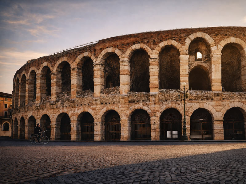 Arena di Verona al tramonto, vista dalla piazza con pavimentazione in ciottoli e una persona in bicicletta davanti agli archi dell’anfiteatro.