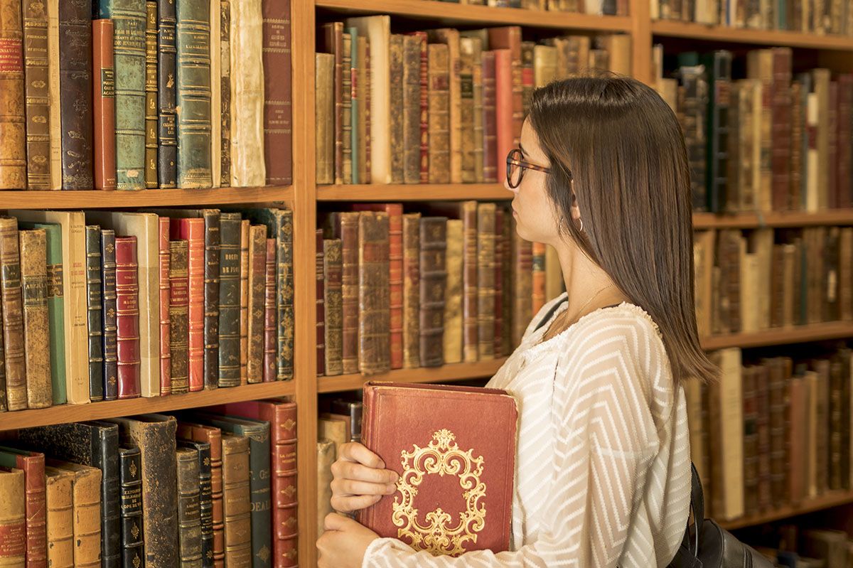 ragazza con libro sotto braccia guarda scaffale di libri antichi all'interno di una biblioteca