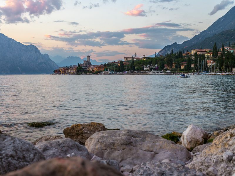 Vista sul Lago di Garda al tramonto, con le rocce in primo piano, il borgo di Malcesine sulla riva e le montagne sullo sfondo.