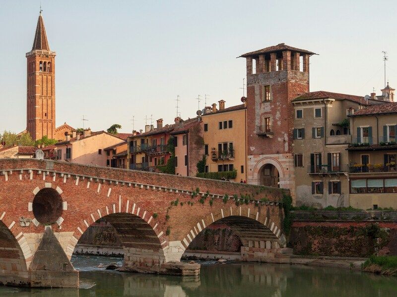 sta sul Ponte Pietra a Verona al tramonto, con il fiume Adige in primo piano, edifici storici affacciati sull’acqua e campanile in mattoni sullo sfondo.