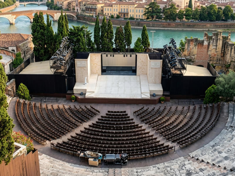 Veduta panoramica dall'alto del Teatro Romano di Verona, che mostra l'antica cavea semicircolare in pietra, il palcoscenico allestito con strutture moderne per spettacoli, il fiume Adige che scorre alle spalle e lo storico Ponte Pietra sullo sfondo.
