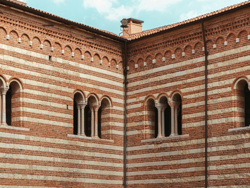 Scorcio architettonico del Cortile del Mercato Vecchio a Verona, caratterizzato da pareti con tipiche fasce alternate di mattoni rossi e tufo bianco.