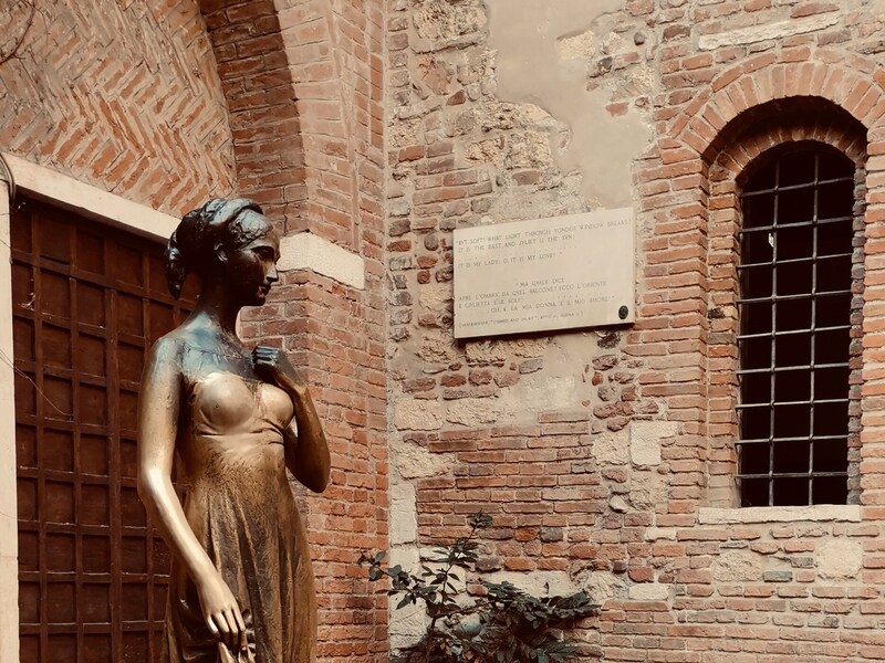 Statua in bronzo di Giulietta nel cortile della Casa di Giulietta a Verona, accanto a una parete storica in mattoni con finestra ad arco.